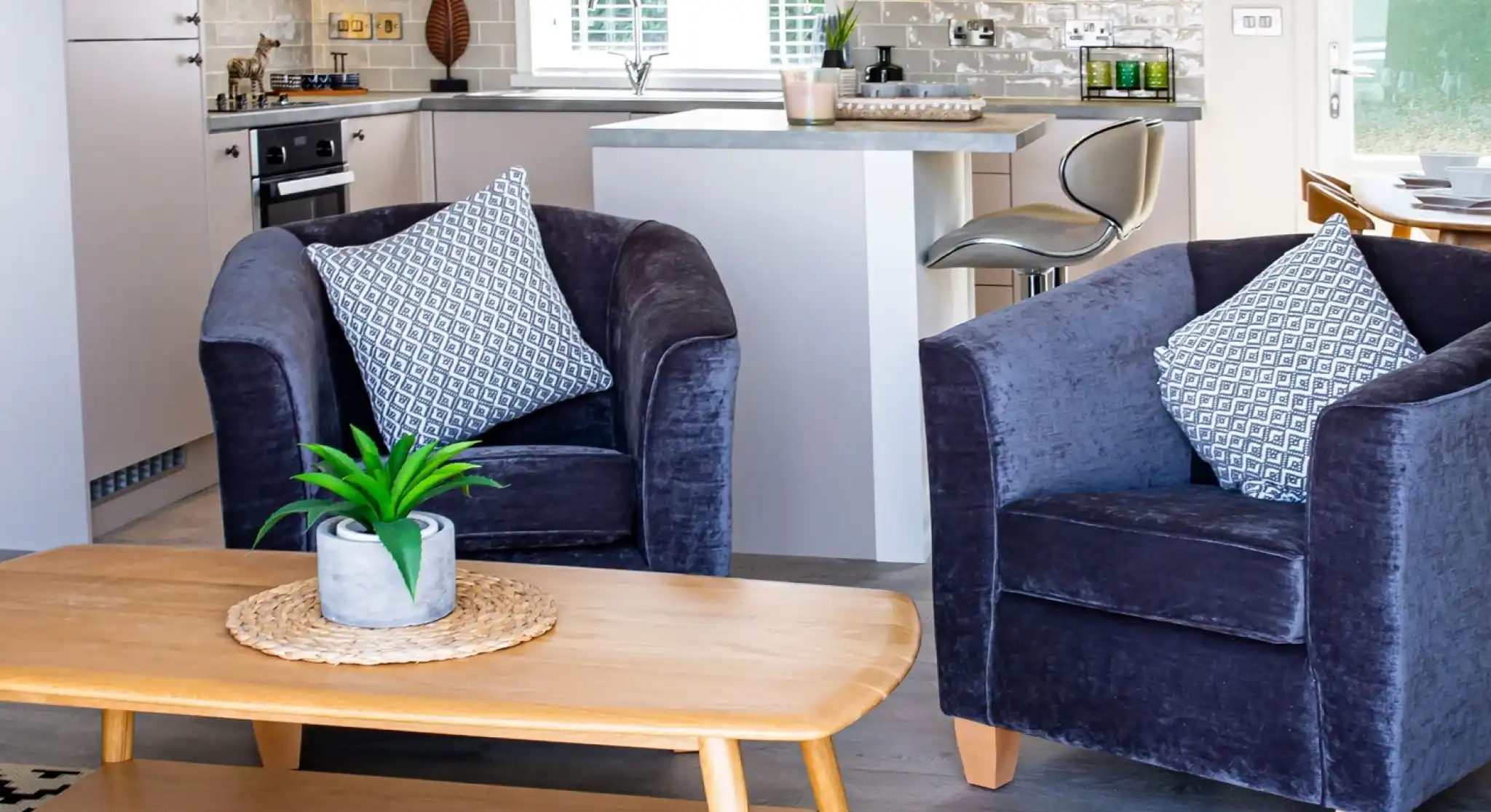Cosy purple velvet tub chairs surrounding a light wooden coffee table with kitchen in the background