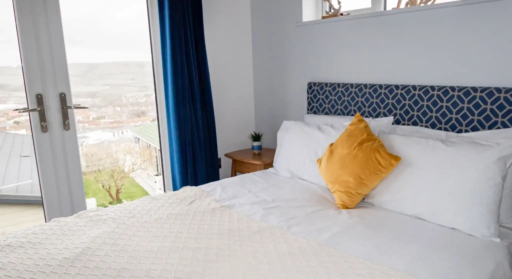 Double bed with white sheets and plush headboard in bedroom with patio doors and views over Swanage Coastal Park