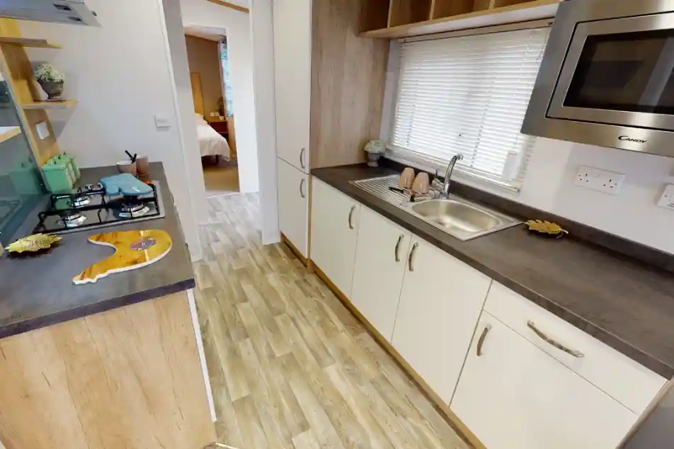 A modern kitchen with light wood cabinetry and a stainless steel sink. The countertop features a gas stove and a microwave overhead. Natural light filters through the window, illuminating the space. In the background, a doorway leads to a bedroom.