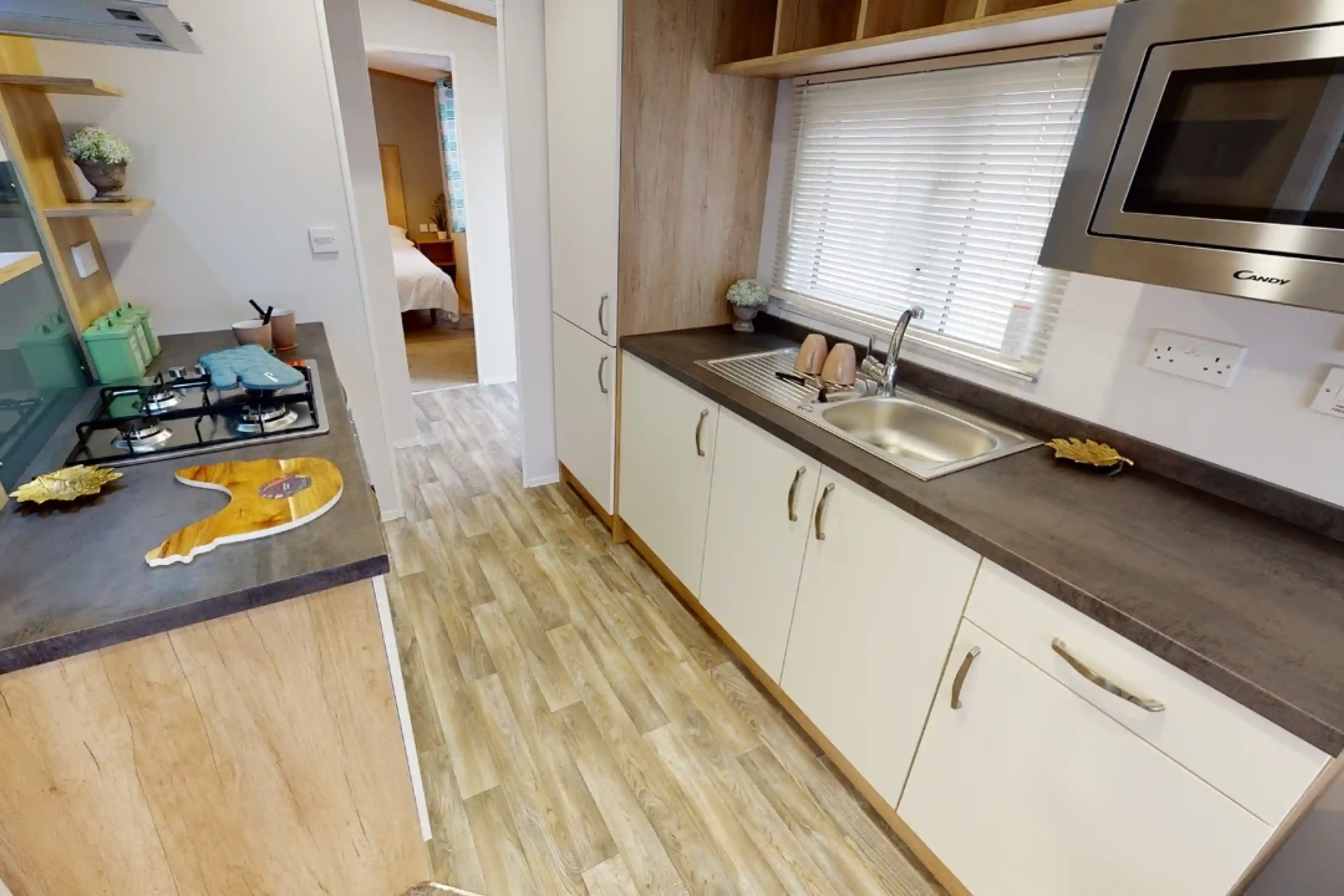 A modern kitchen with light wood cabinetry and a stainless steel sink. The countertop features a gas stove and a microwave overhead. Natural light filters through the window, illuminating the space. In the background, a doorway leads to a bedroom.