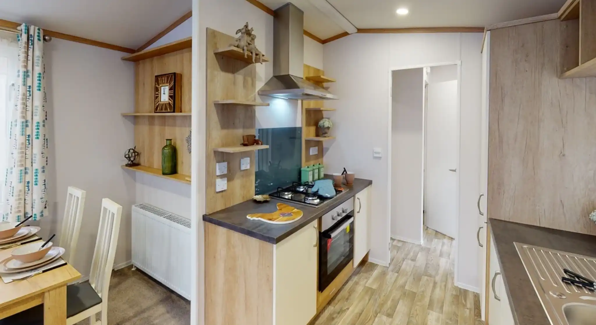 A modern kitchen featuring light wooden cabinetry, a gas stove, and an extractor hood. Open shelves display decorative items and kitchenware. A dining area with a small table and white chairs is visible. The flooring is a light wood laminate.
