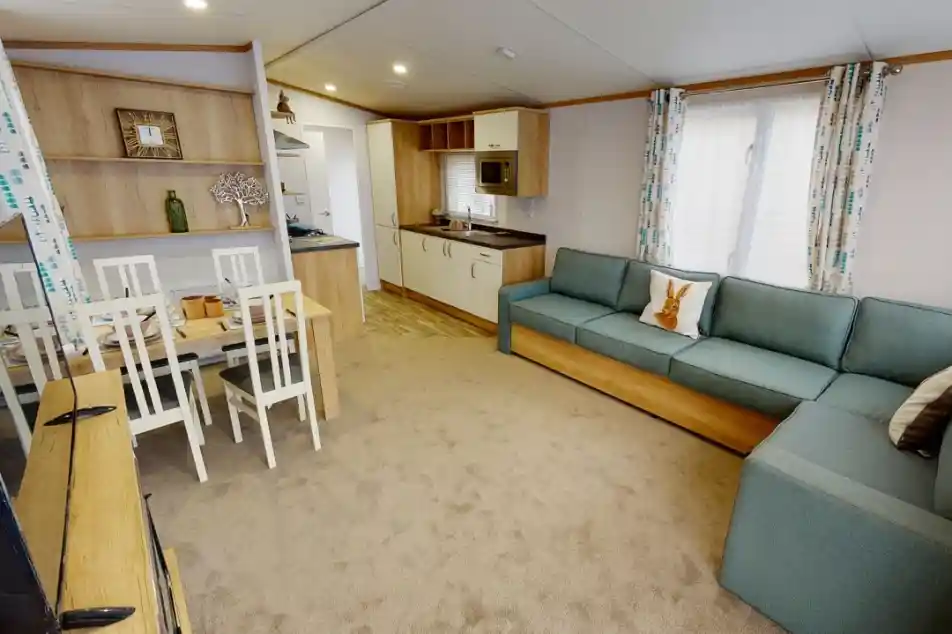 A cozy living space featuring a light-colored carpet, a blue sectional sofa, and a wooden dining table with white chairs. The kitchen area includes modern appliances and wooden cabinetry, illuminated by natural light through a window with patterned curtains.