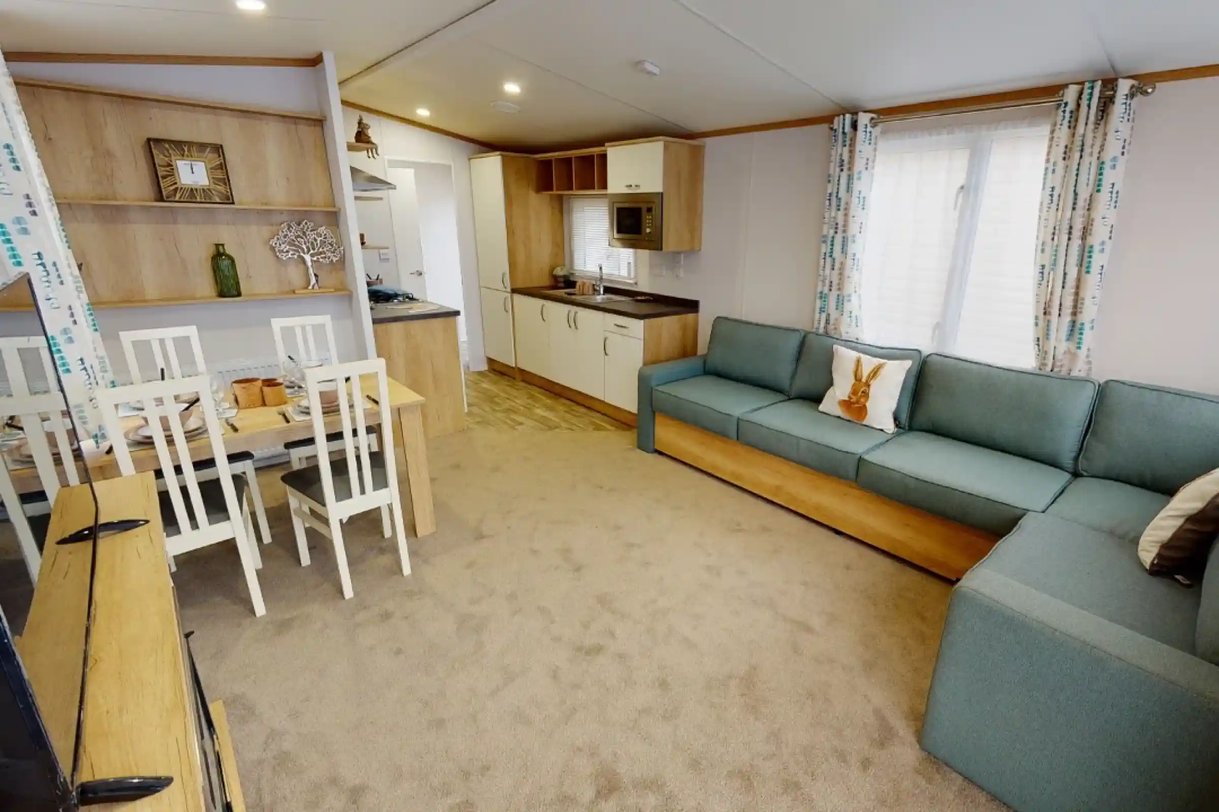 A cozy living space featuring a light-colored carpet, a blue sectional sofa, and a wooden dining table with white chairs. The kitchen area includes modern appliances and wooden cabinetry, illuminated by natural light through a window with patterned curtains.