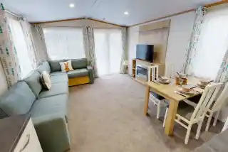 A cozy living area featuring a light blue sectional sofa, a wooden dining table set for four, and a large TV mounted on the wall. Natural light streams in through large windows with patterned curtains. The carpet is beige, creating a warm atmosphere.