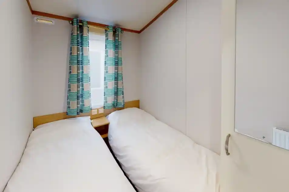 Cozy bedroom with two twin beds positioned side by side, light bedding, and colorful patterned curtains. A window allows natural light, and a mirror is visible on the door.