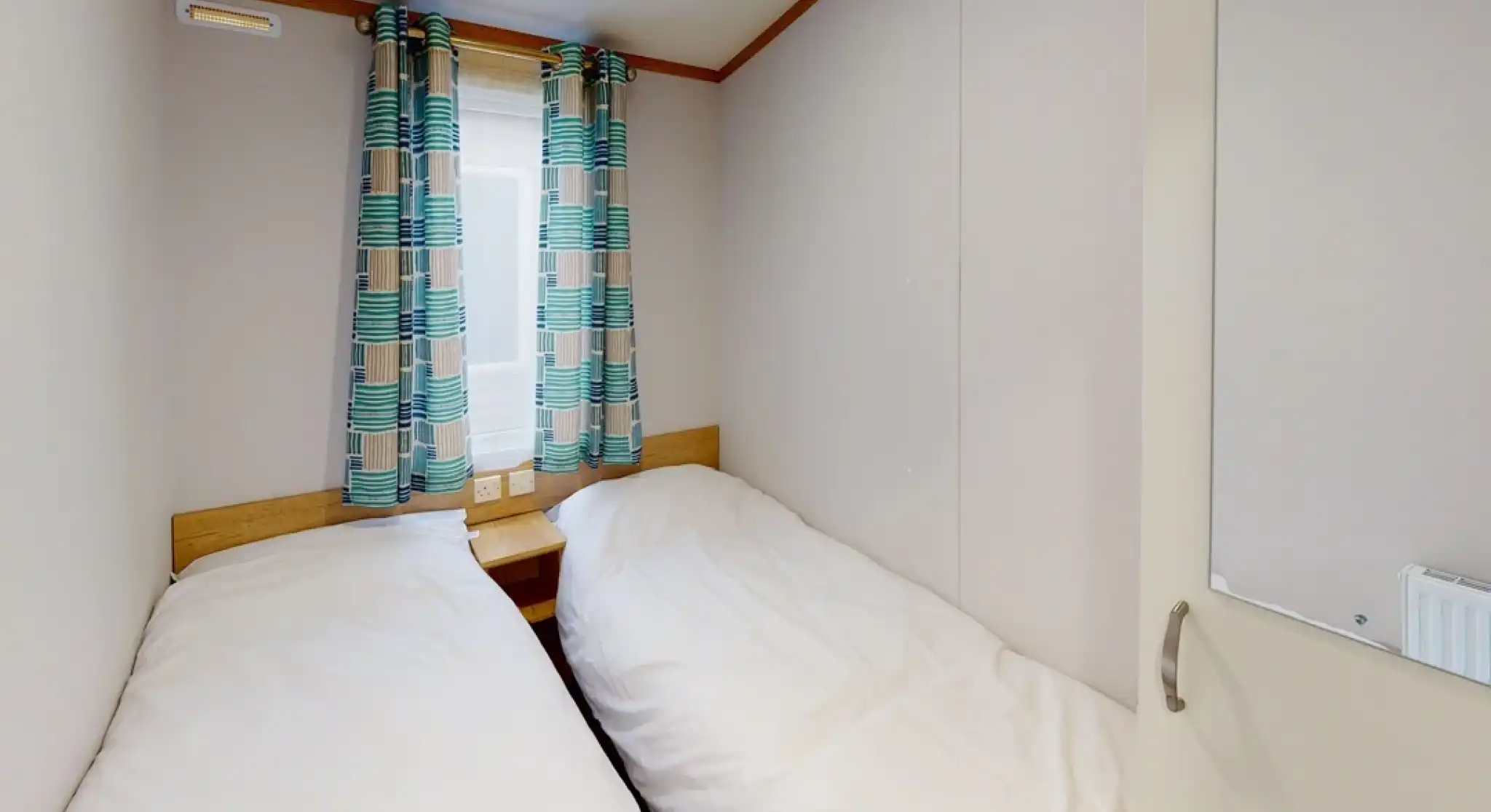 Cozy bedroom with two twin beds positioned side by side, light bedding, and colorful patterned curtains. A window allows natural light, and a mirror is visible on the door.