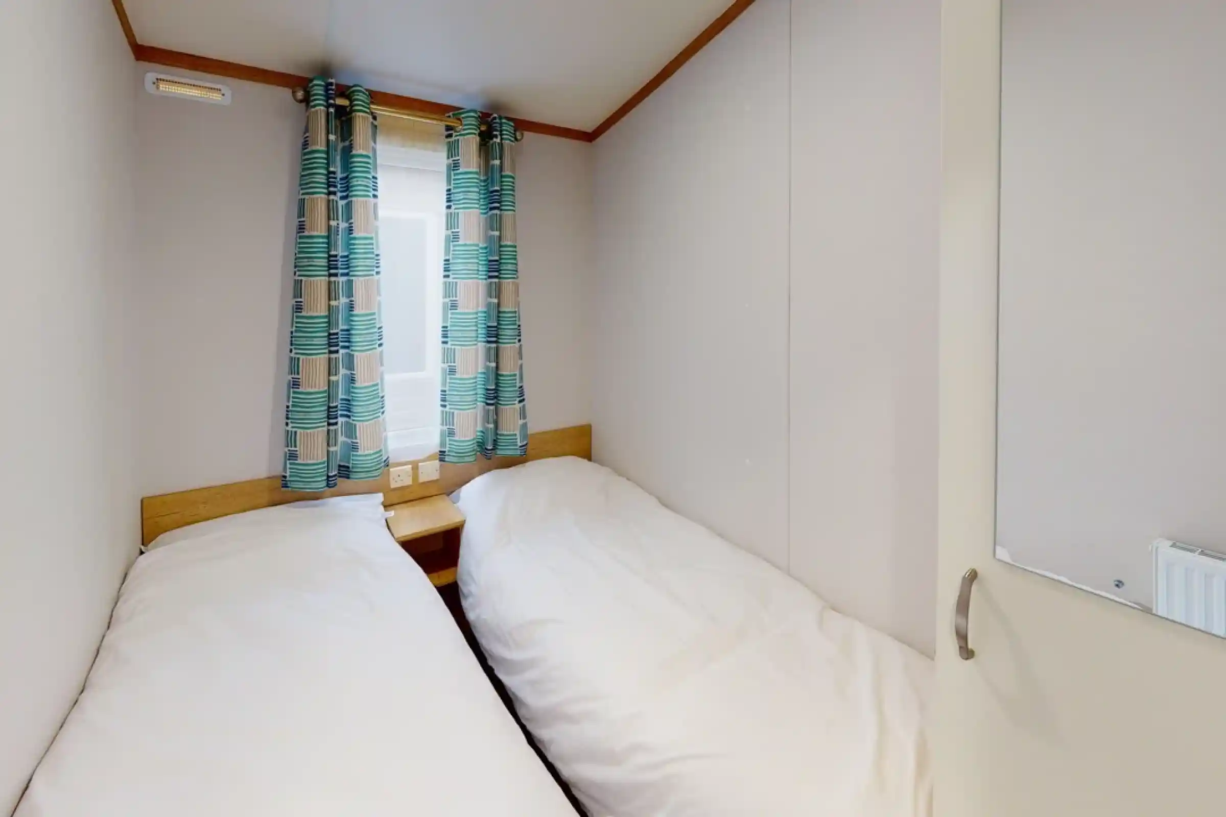 Cozy bedroom with two twin beds positioned side by side, light bedding, and colorful patterned curtains. A window allows natural light, and a mirror is visible on the door.