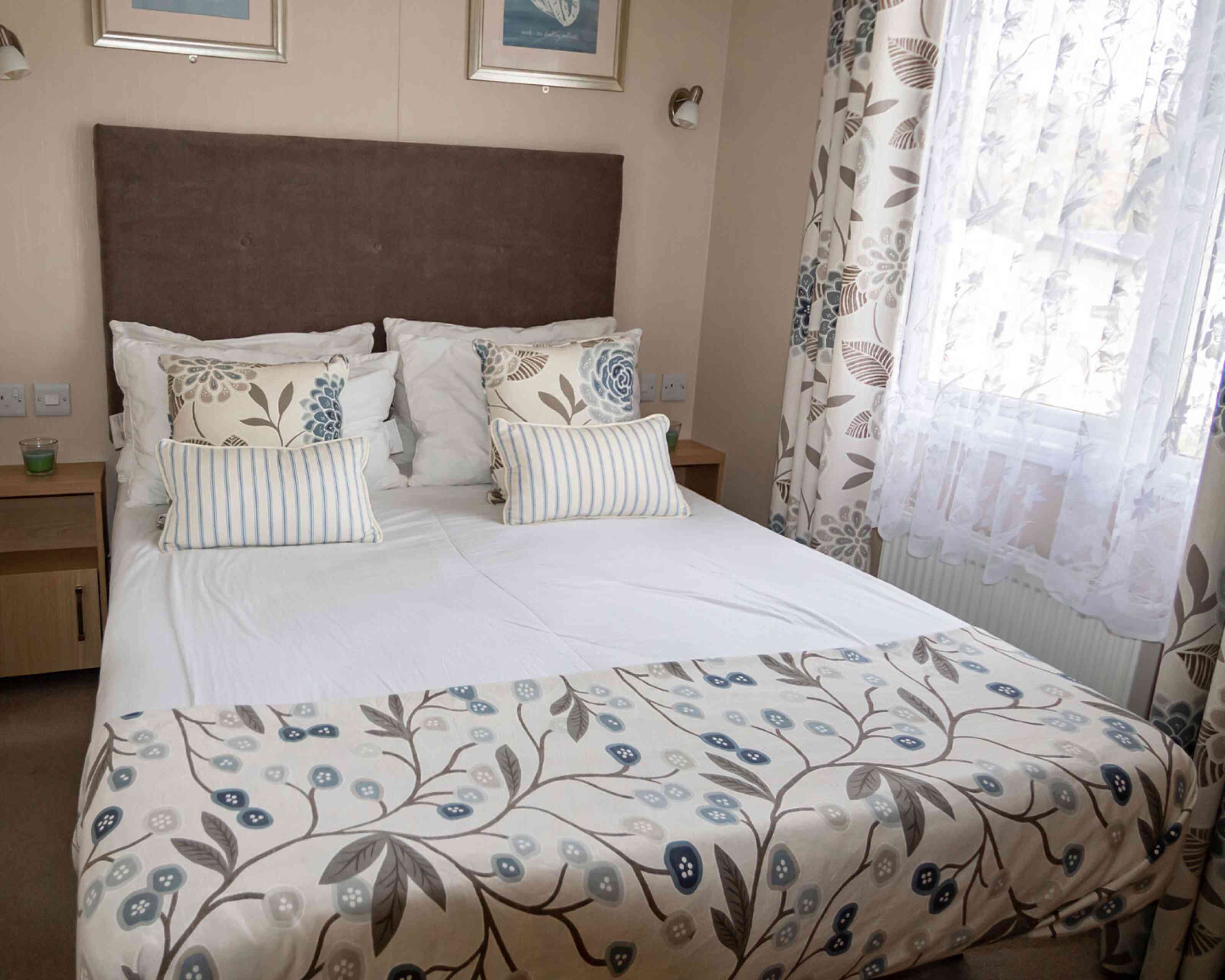 Cosy bedroom with a large bed featuring patterned floral bedding and pillows. Soft, natural light filters through white lace curtains, creating a calm ambiance.