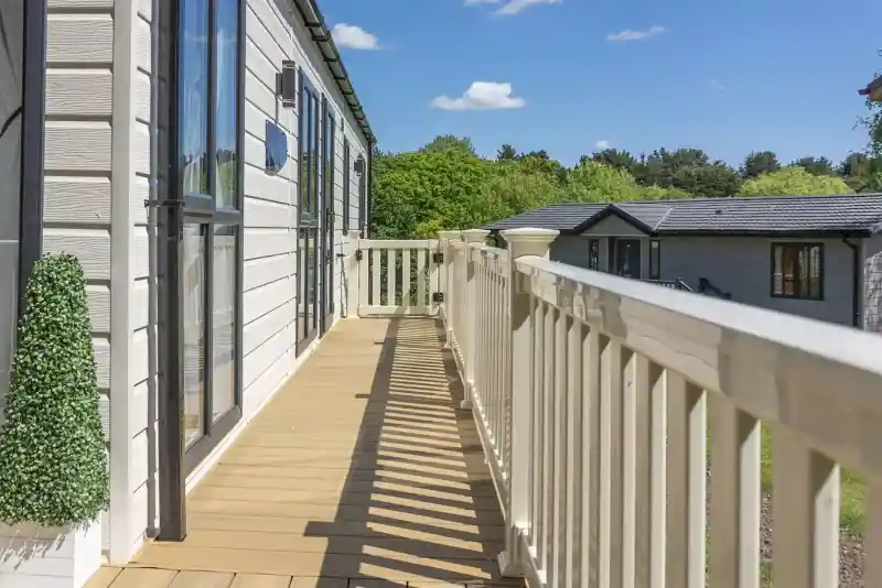 A wooden balcony with a railing overlooks a green landscape, featuring trees and neighbouring buildings under a clear blue sky with scattered clouds.