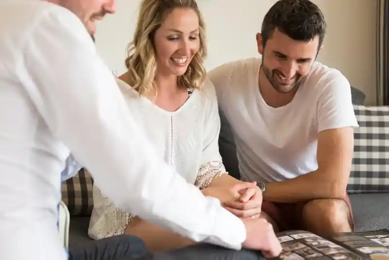 Three people sit closely together on a couch, looking at a photo album. One person gestures towards the album while the others smile, engaged in conversation. The background features a cosy living space with decorative cushions.