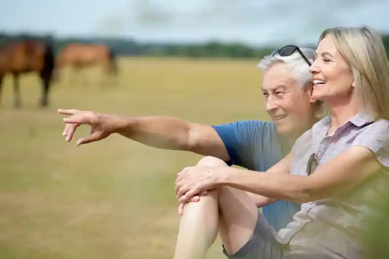 An older couple sits together in a grassy field, smiling and enjoying each other's company. The man points out something in the distance, while the woman looks happily at him. Several horses graze in the background under a clear sky.