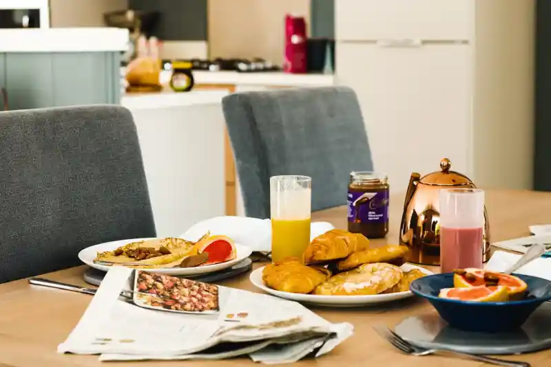 A cosy breakfast setup on a wooden table featuring croissants, pancakes, fruit, juice, and a newspaper. Two grey chairs are positioned around the table, with a modern kitchen visible in the background.