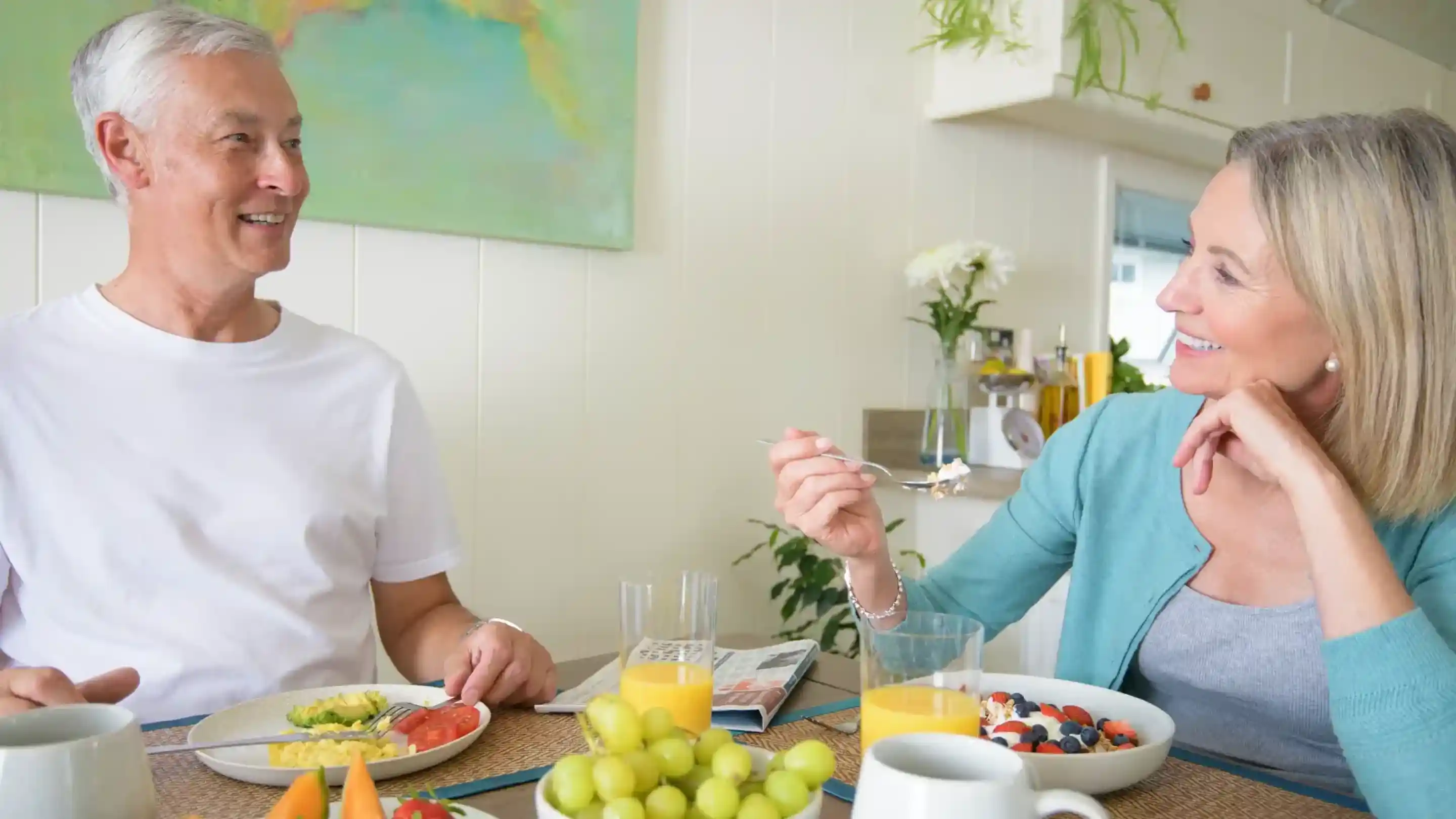 A man and woman enjoy breakfast together at a kitchen table, smiling and engaging in conversation. The table is adorned with colourful plates of food, including fruit and eggs, along with glasses of juice. A vibrant painting hangs in the background.