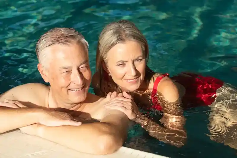 A happy older couple enjoying time together by the edge of a swimming pool, with the woman gently resting her arm on the man's shoulder. They both have bright smiles and appear relaxed.