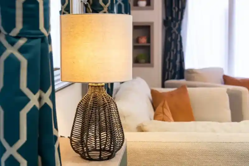 A stylish woven lamp with a warm glow sits on a table next to a cosy sofa, framed by patterned curtains and a softly lit room.