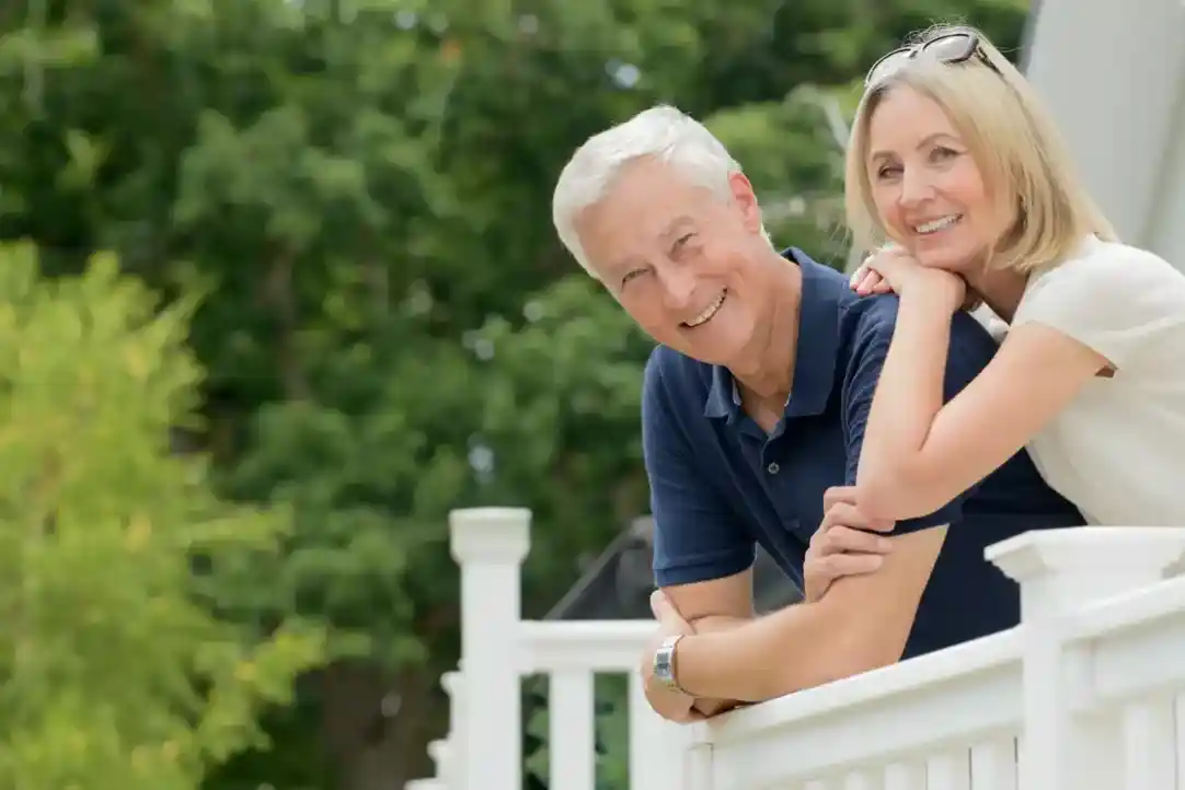 A smiling couple stands closely together on a white railing, surrounded by lush greenery. The man has short grey hair and is wearing a navy polo shirt, while the woman has blonde hair and is dressed in a light top.