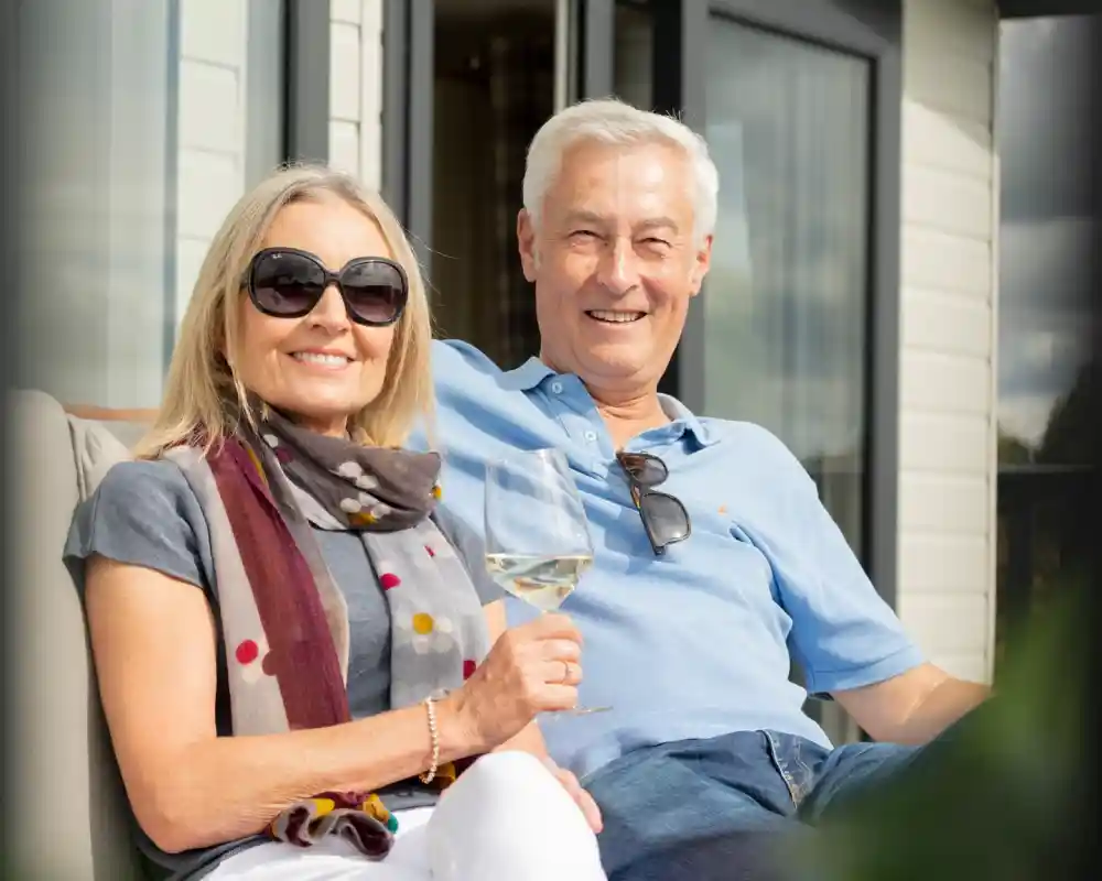 A smiling woman with sunglasses and a scarf sits next to a man in a blue polo shirt, both enjoying a sunny day outdoors. She holds a glass of white wine while they relax on a couch.