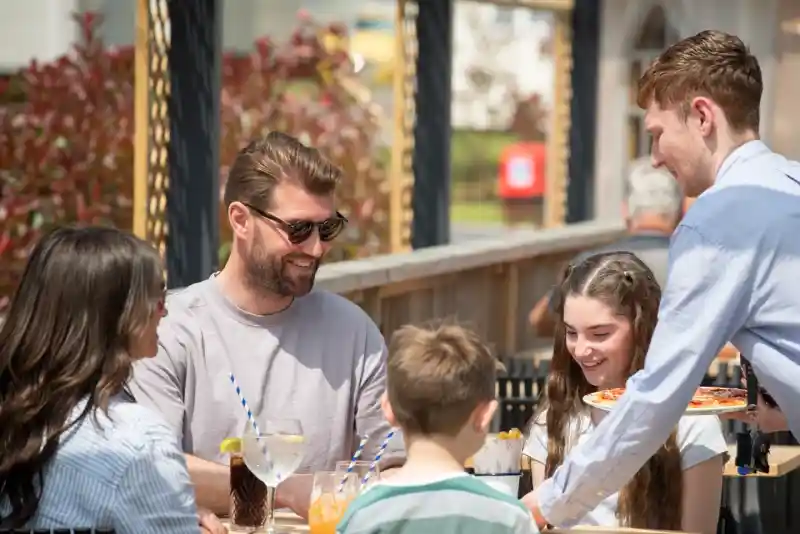 A group of four people sits at an outdoor table enjoying a meal. Two adults smile at each other, while a boy and a girl sit across from them. A server approaches the table with a pizza. Drinks, including a cocktail and soda, are on the table with a sunny backdrop.