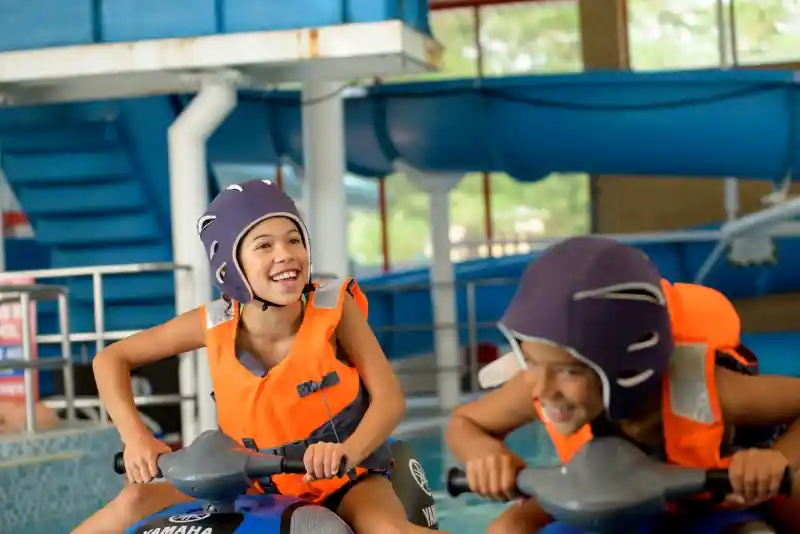 Two children joyfully ride jet skis in a swimming pool, wearing life jackets and helmets. They are smiling and having fun, surrounded by blue water and a playful indoor environment.