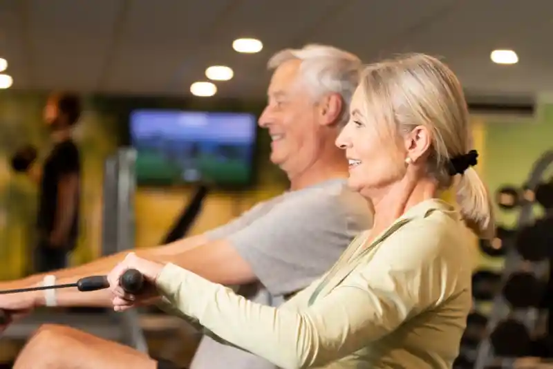 An older man and woman are exercising together on rowing machines in a gym. Both are smiling and appear focused, with the woman wearing a light green jacket and the man in a grey t-shirt. In the background, other gym equipment and people can be seen.
