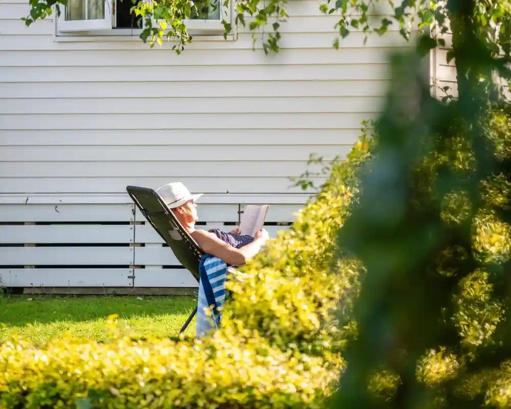 A person relaxes in a lounge chair in a sunny garden, wearing a straw hat and reading a book. Green bushes frame the scene, with a white house in the background.