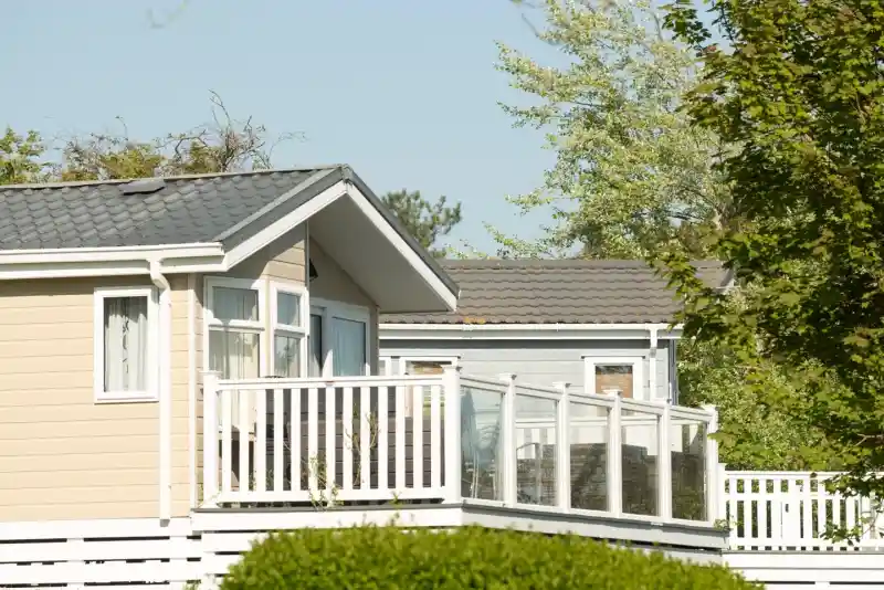 Two modern holiday cottages with wooden decks, surrounded by lush greenery and trees under a clear blue sky.