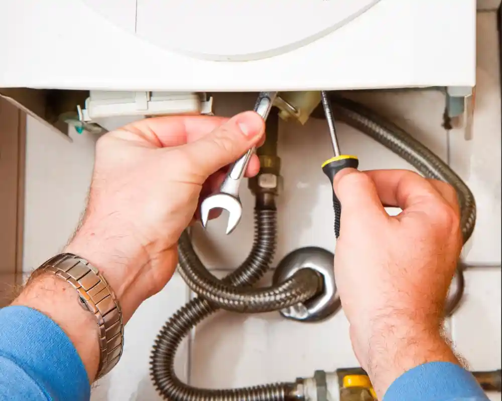 Hands holding a wrench and screwdriver while working on plumbing under a sink. Various pipes and fittings are visible.