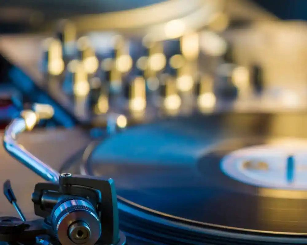 A close-up of a vinyl record on a turntable with a tonearm, featuring a blurred background of knobs and controls.