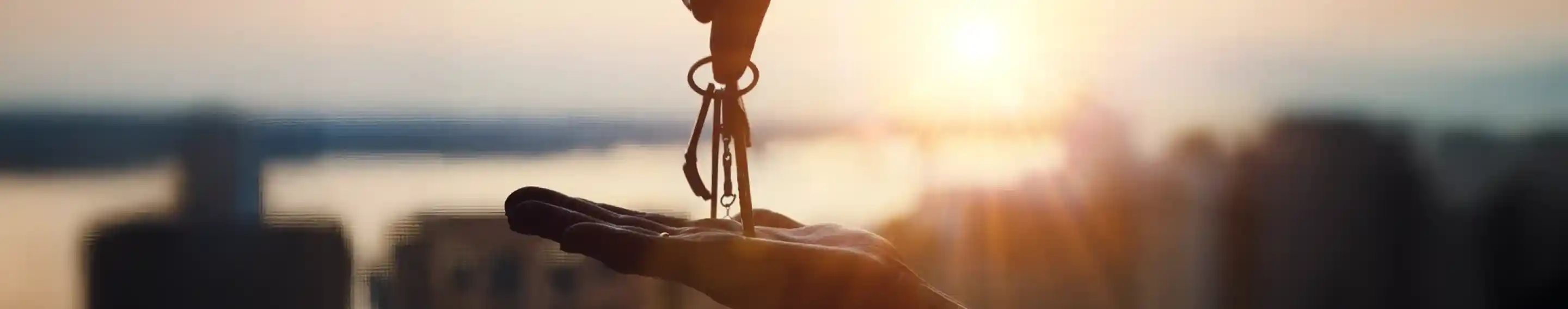 A hand is passing a set of keys to another hand, with a sunset in the background illuminating the scene. The silhouettes of buildings are visible below, creating a sense of urban life.
