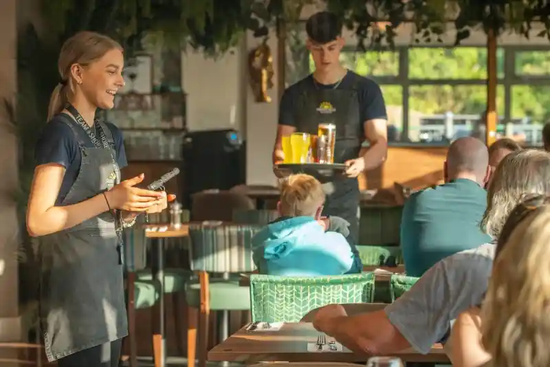 A young female server smiles while holding a notepad, standing in a vibrant restaurant. In the background, a male server carries a tray with drinks, while patrons enjoy their meals at the tables. Lush greenery decorates the space.