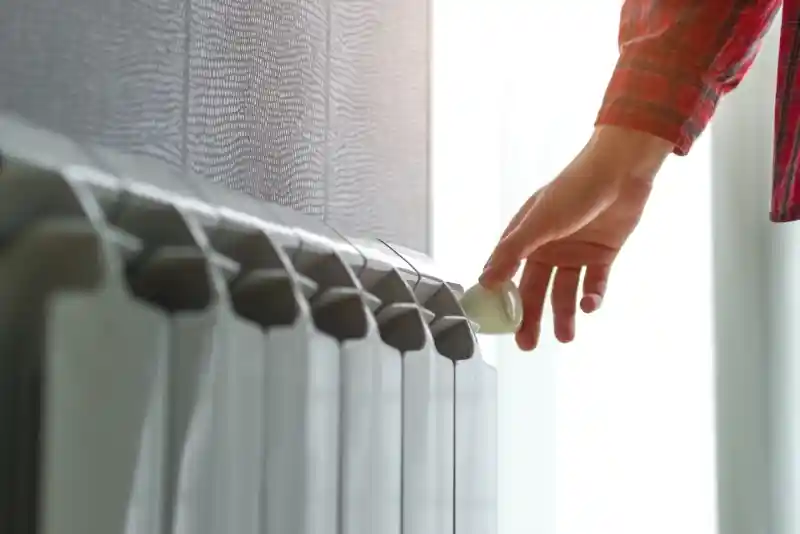 A person in a checked shirt adjusts the temperature dial on a radiator, with sunlight streaming in through a nearby window.