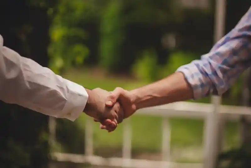 Two hands are shaking in a handshake outdoors, with greenery blurred in the background. One hand is wearing a white long-sleeve shirt, and the other is wearing a checked shirt.