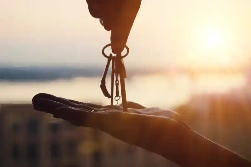 A hand is reaching out to give a set of keys into another open palm, with a sunset in the background creating a warm glow.