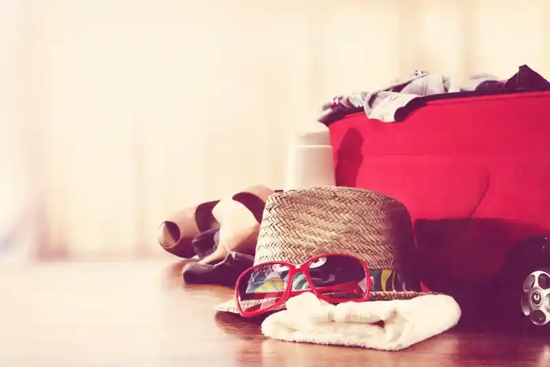 A straw hat with colourful band, red sunglasses, and a white towel are placed on a wooden table next to a red suitcase, with various clothing items scattered around. The background is softly blurred, suggesting a warm, vacation vibe.