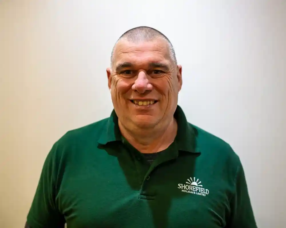 A smiling man with a closely cropped haircut, wearing a dark green polo shirt with the logo "SHOREFIELD." He stands against a plain light-colored background.