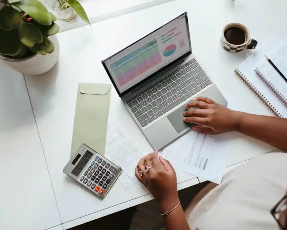 A person is working at a white desk, using a laptop to review spreadsheets. Nearby, a calculator rests on financial documents, and a steaming cup of coffee is visible. A green envelope and a notepad with notes are also on the desk.