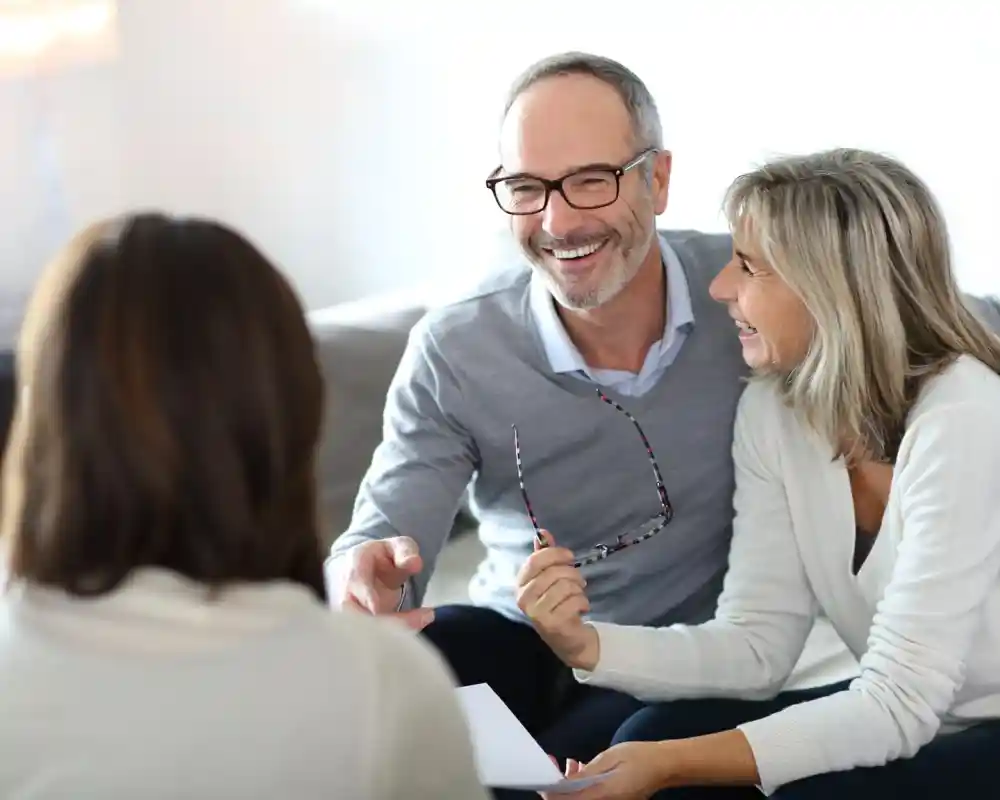 A smiling middle-aged man and woman engage in a conversation with another person, sitting together in a cosy living room. The man holds a pair of glasses, while the woman looks happily at him. All three appear to be in a relaxed and positive atmosphere.