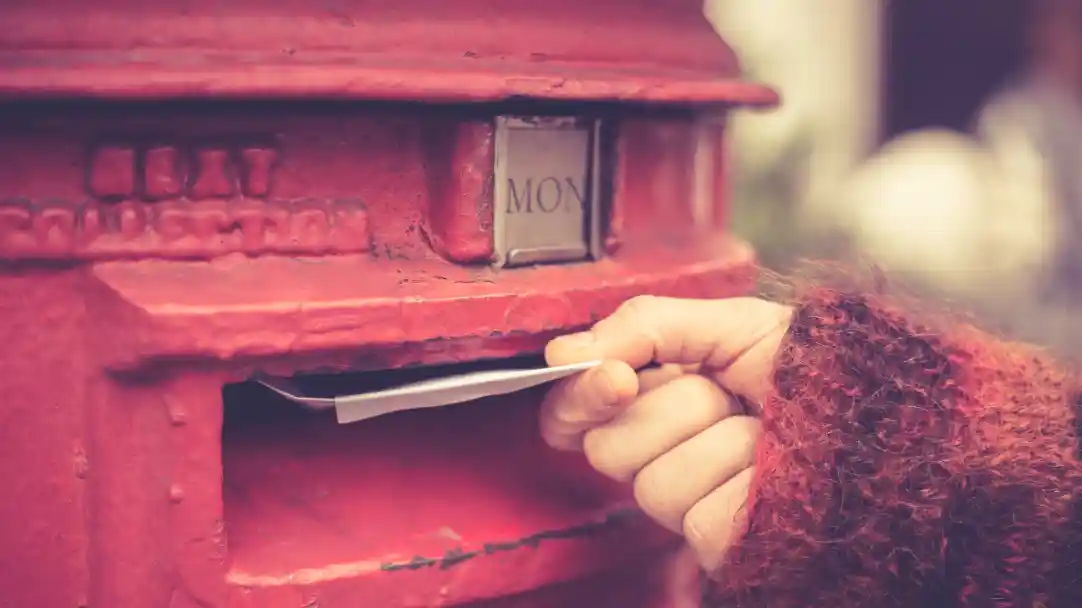 A hand in a fuzzy sweater is inserting a letter into a vintage red mailbox with the label "MON" on it.