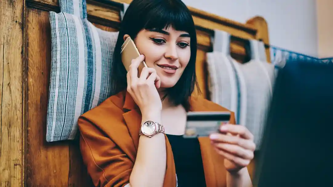 A young woman with dark hair and bangs smiles while holding a credit card in one hand and talking on her phone with the other, sitting in a cosy setting with striped cushions in the background.