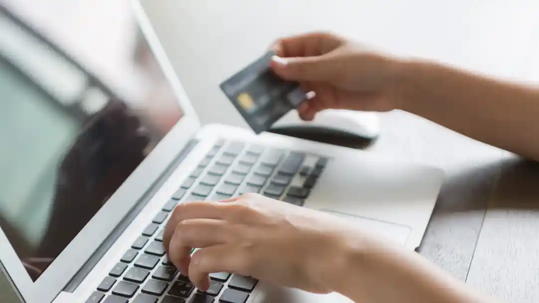 A person typing on a laptop keyboard while holding a credit card in their right hand. The laptop screen shows a reflection of the individual.