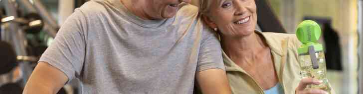 A smiling older couple sits together in a gym, with the woman holding a green water bottle. They appear to be enjoying each other's company after a workout. The gym features exercise equipment in the background.
