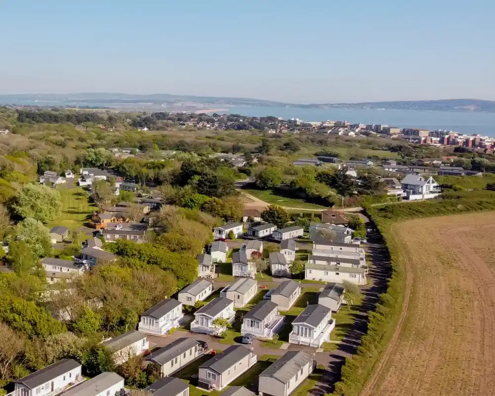 Aerial view of a residential area featuring rows of mobile homes surrounded by greenery, with a distant coastal view and hills visible in the background.
