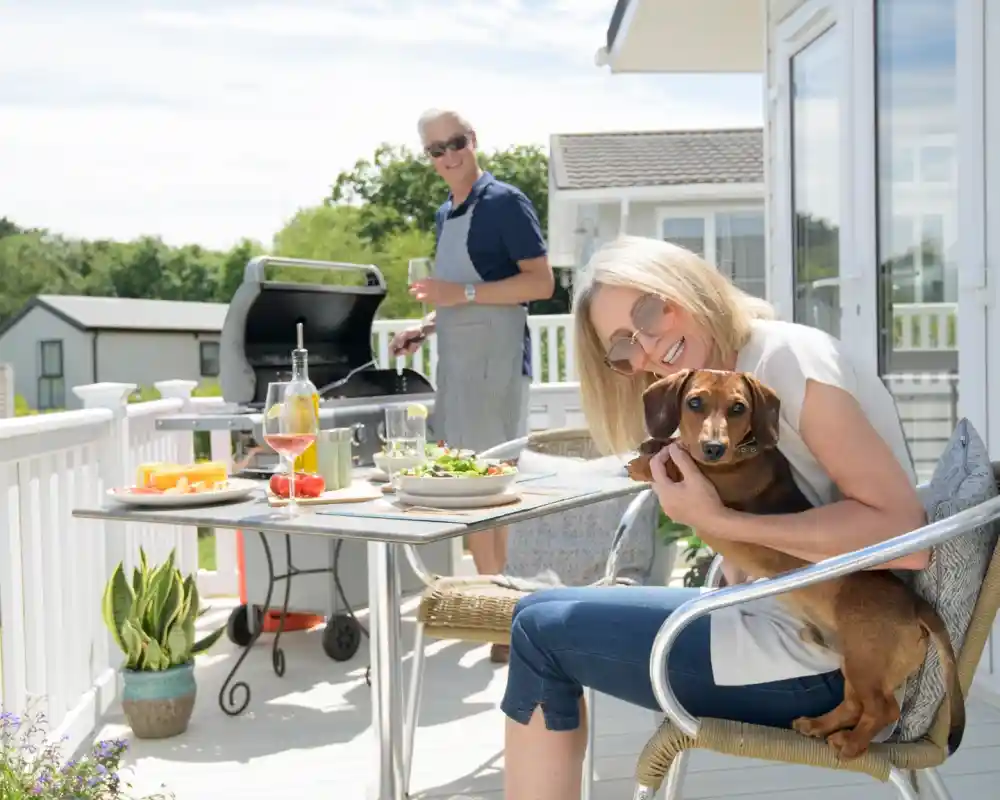 A woman sitting at an outdoor table, holding a small dog and smiling. In the background, a man stands by a grill, preparing food, with a glass in hand. The setting is bright and cheerful, with greenery and a sunny sky.