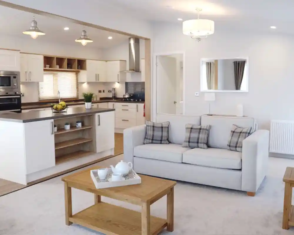 A modern, open-plan living area featuring a light gray sofa with plaid cushions, a wooden coffee table, and a cozy carpet. The kitchen is visible in the background, showcasing white cabinets and stainless-steel appliances. Soft lighting illuminates the space, creating a warm and inviting atmosphere.
