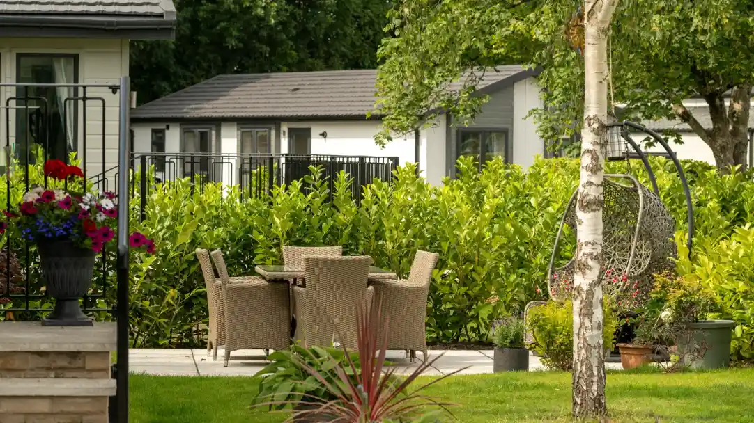 A cozy outdoor patio with a wicker dining set surrounded by lush green hedges. In the background, there are modern cabins and a decorative hanging chair. Colorful potted flowers enhance the serene garden atmosphere.