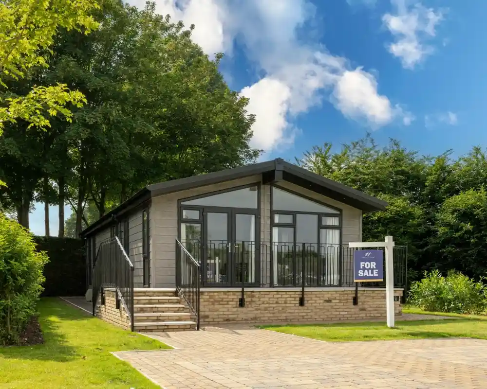 A modern, single-story home with large windows and a black roof is shown for sale. It features a well-kept lawn, a paved driveway, and surrounding greenery under a bright blue sky with fluffy clouds.