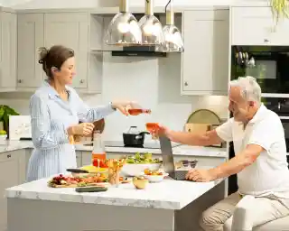 A woman in a blue striped shirt pours a drink into a glass for a man sitting at a kitchen island. The island is adorned with various food items, and there are pendant lights hanging above, creating a cozy atmosphere. The man is using a laptop and smiling at the woman.