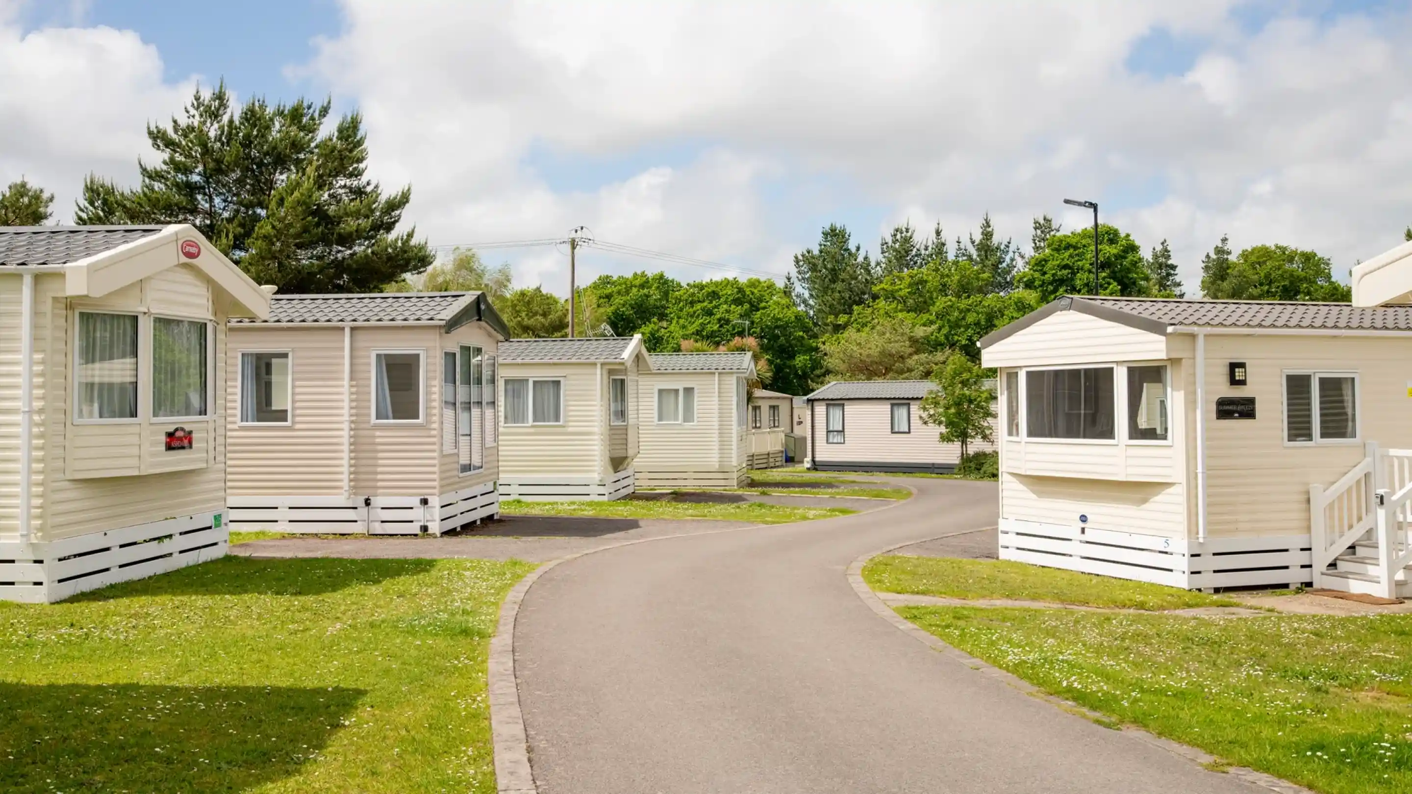 A peaceful holiday park with several static caravans lining a winding road. Lush green grass and trees are visible on either side, under a partly cloudy sky.