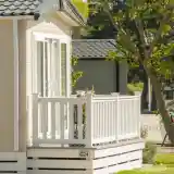 A light-colored cabin with a small white balcony, surrounded by greenery and a tree. The cabin features large windows and a modern design.