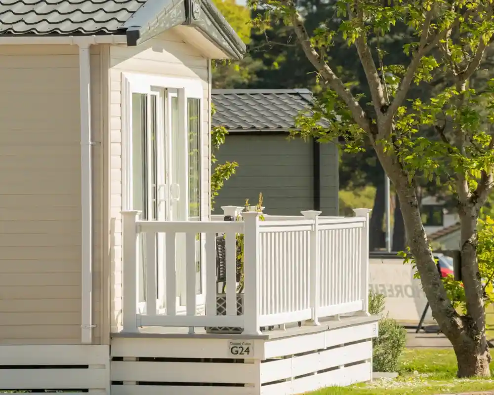 A light-colored cabin with a small white balcony, surrounded by greenery and a tree. The cabin features large windows and a modern design.